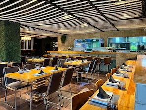 A restaurant with multiple tables and chairs, a bar area, and a kitchen visible in the background. - Hotel GH Guaparo Inn Valencia (Naguanagua)