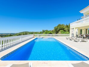 Une piscine dotée d’une balustrade blanche, un espace détente couvert avec des parasols, et une vue sur le paysage.