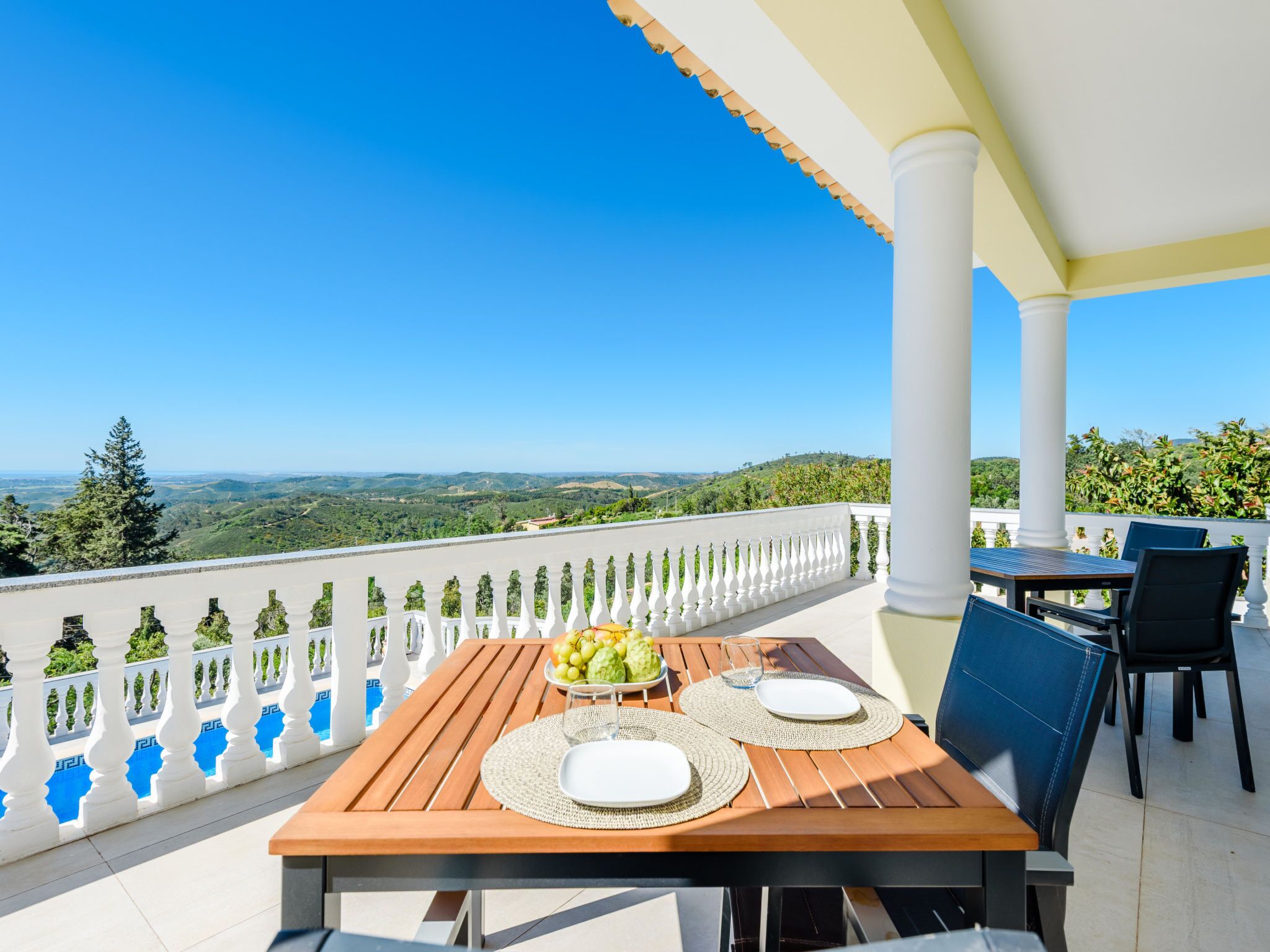 A wooden table set with plates, a bowl of fruit, and a glass of water on a balcony with a white balustrade and a view of the landscape.