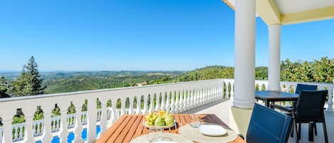 A wooden table set with plates, a bowl of fruit, and a glass of water on a balcony with a white balustrade and a view of the landscape.