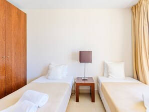 Ein Hotelzimmer mit zwei Betten, einem Holzschrank, einem Nachttisch mit Lampe sowie einem Fenster mit Vorhängen.