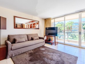 Ein modernes Wohnzimmer mit einer Couch, einem Flachbildfernseher und einer großen Schiebeglastür, die auf einen Balkon mit Blick auf Palmen führt.