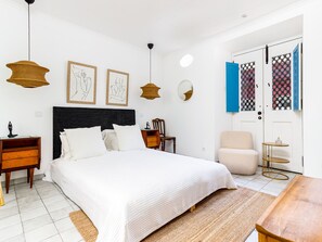 Ein Schlafzimmer mit einem großen Bett, Nachttischen, einem Sessel, einem kleinen Tisch und einer Tür mit blauem Rollo.