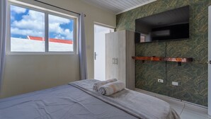 A bedroom with a bed, a wardrobe, a TV mounted on the wall, and a window with a view of the sky. - Aruba Vibe Apartments #2 | A charming place near the beaches and the city center (Oranjestad)
