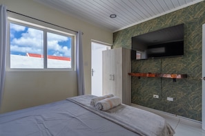 A bedroom with a bed, a wardrobe, a TV mounted on the wall, and a window with a view of the sky. - Aruba Vibe Apartments #2 | A charming place near the beaches and the city center (Oranjestad)