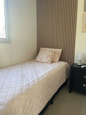 Ein Bett mit einer gesteppten weißen Bettdecke, einem floralen Kopfkissen und einem schlichten Kopfkissen, vor einer Wand mit vertikalen Jalousien.