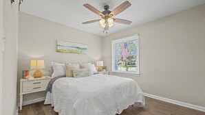 Ein Schlafzimmer mit einem Bett, Nachttischen, einem Deckenventilator, einem Fenster mit Jalousien und einem Bild an der Wand.
