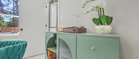 Ein Zimmer mit einem türkisfarbenen Sessel, einem Vintage-Radio, einer Zierpflanze und einem Fenster mit Blick auf Bäume.