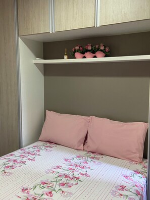 Un dormitorio con cama, dos almohadas, repisas con adornos y una colcha estampada con motivos florales.