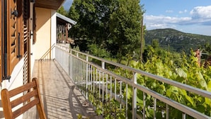 Ein Balkon mit einem Holzbalken, einem Stuhl und einem Tisch, mit Blick auf einen Weinberg und eine gebirgige Landschaft.