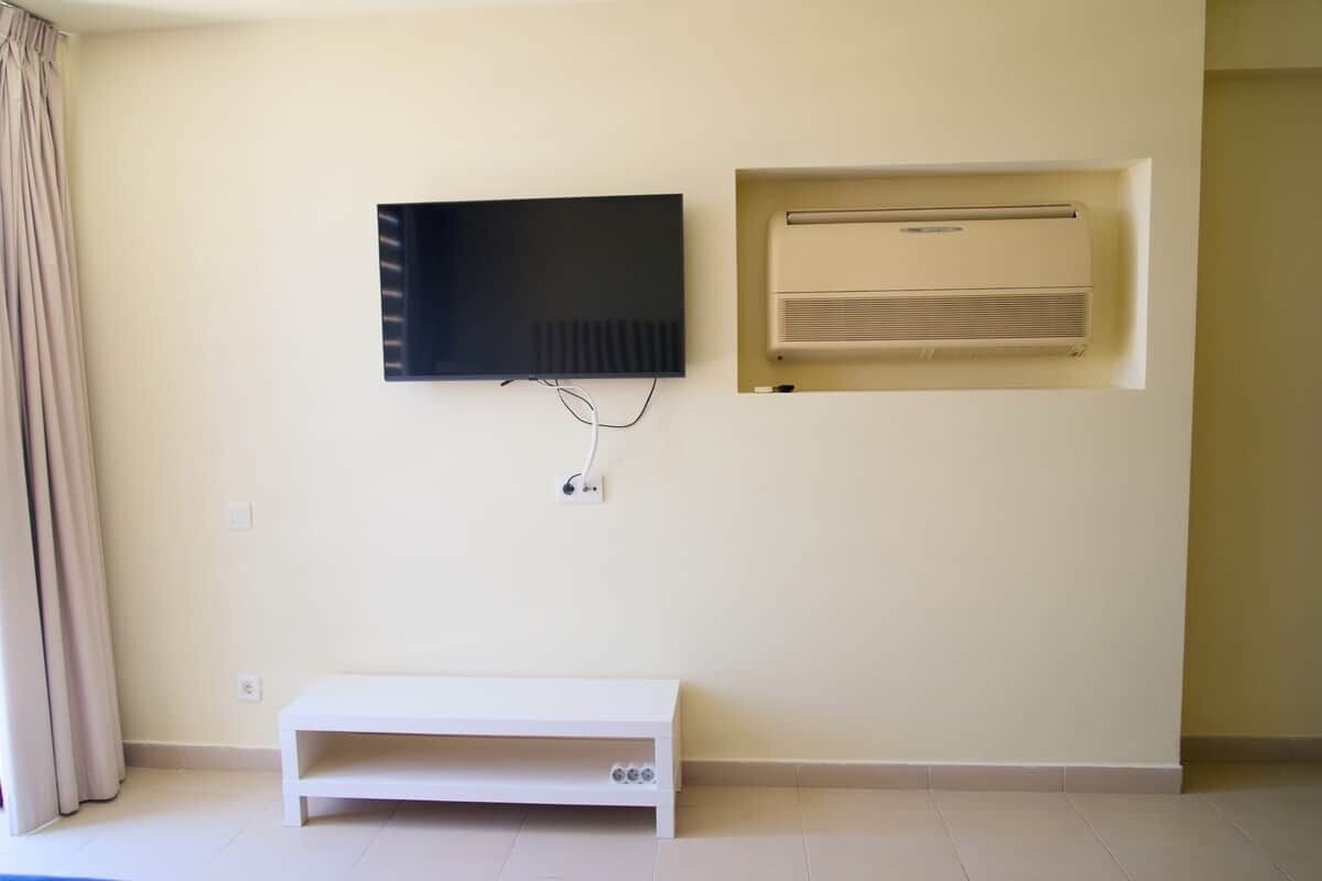 A room with a flat-screen TV mounted on the wall, a built-in air conditioning unit, and a white bench.