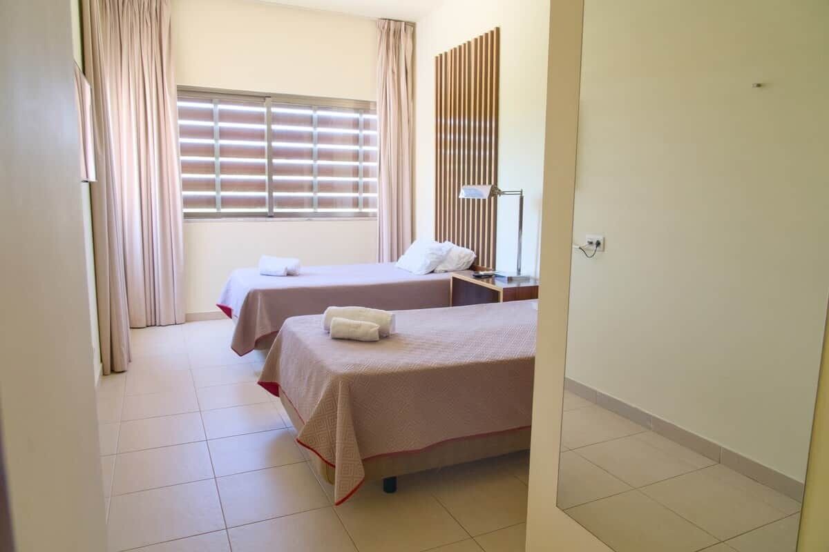 A hotel room with two beds, a window with blinds, a bedside table with a lamp, and a mirror on the wall.