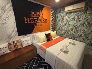 A hotel room with a bed, pillows, a blanket, a wall-mounted air conditioner, and a floral wallpaper.