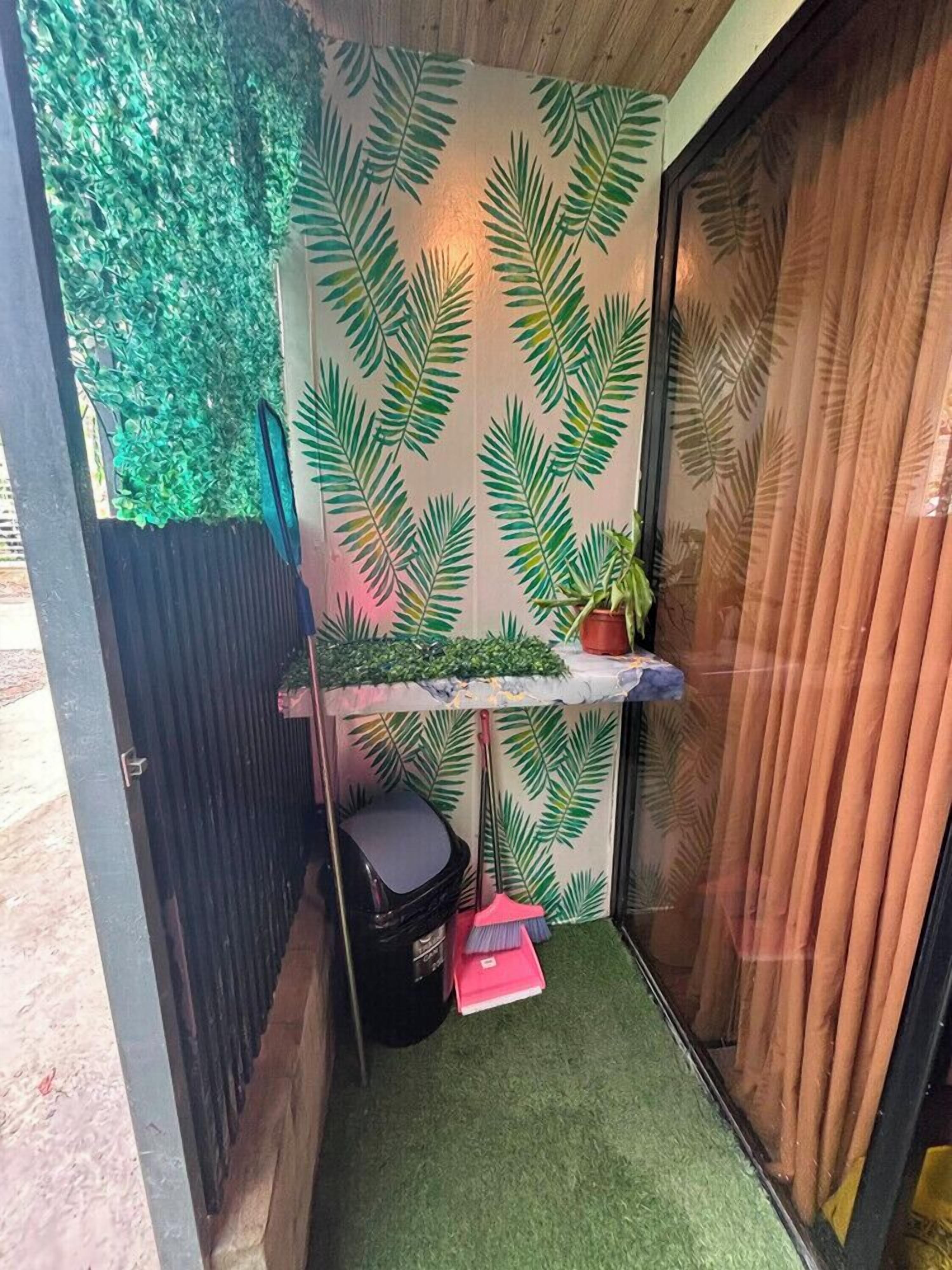 Un espacio reducido con una pared decorada con motivos de hojas verdes, un estante pequeño con una planta en maceta y una escoba apoyada contra la pared.