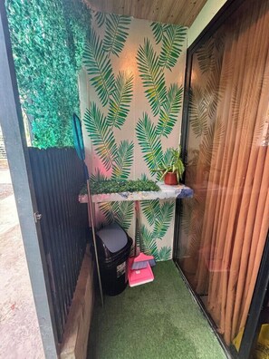 A narrow space with a wall decorated with green leaf patterns, a small shelf with a potted plant, and a broom leaning against the wall.