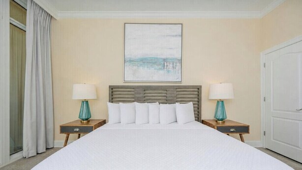 A hotel room with a large bed, two bedside tables with lamps, a headboard, and a painting on the wall. - Phoenix Orange Beach 1202 (Orange Beach)