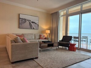 A living room with a sofa, armchair, and a coffee table. There is a painting on the wall and a view through large windows. - Phoenix Orange Beach 1202 (Orange Beach)