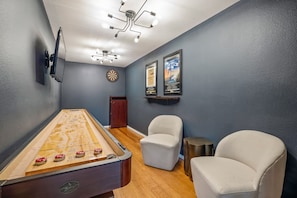 Ein Zimmer mit Shuffleboard-Tisch, zwei Stühlen, Dartscheibe und Wandbildern.