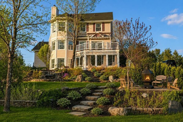 A two-story house with a stone staircase leading to a deck, surrounded by a well-maintained garden with various plants and a fire pit.