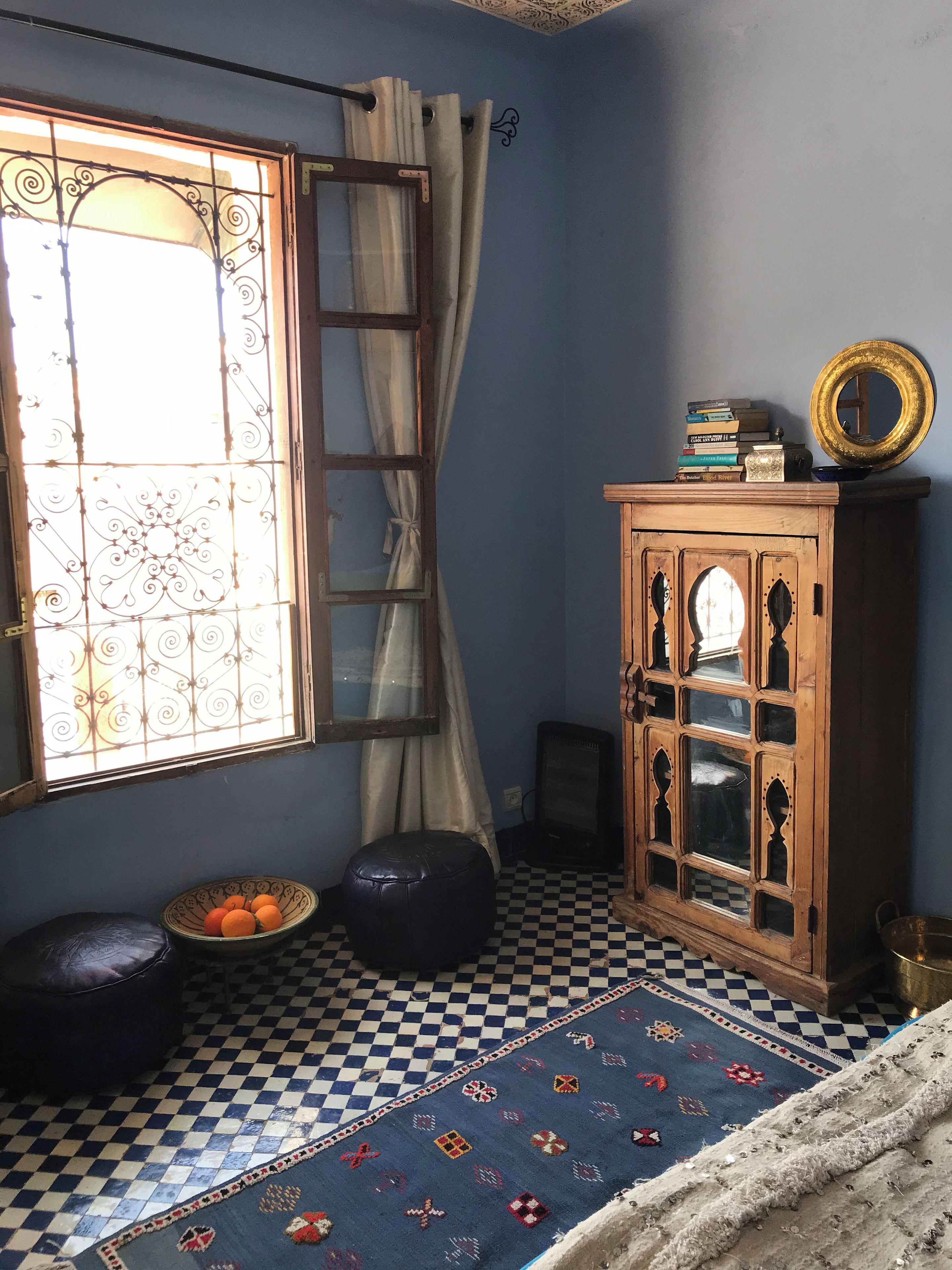 Une pièce avec un sol à carreaux, une fenêtre avec des rideaux, un meuble avec des portes en verre décoratives et un petit tabouret sur lequel sont posées des oranges.
