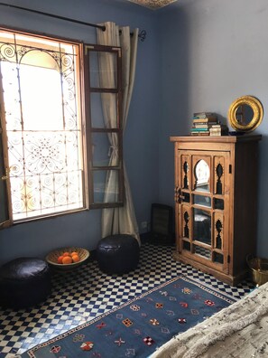 Une pièce avec un sol à carreaux, une fenêtre avec des rideaux, un meuble avec des portes en verre décoratives et un petit tabouret sur lequel sont posées des oranges.