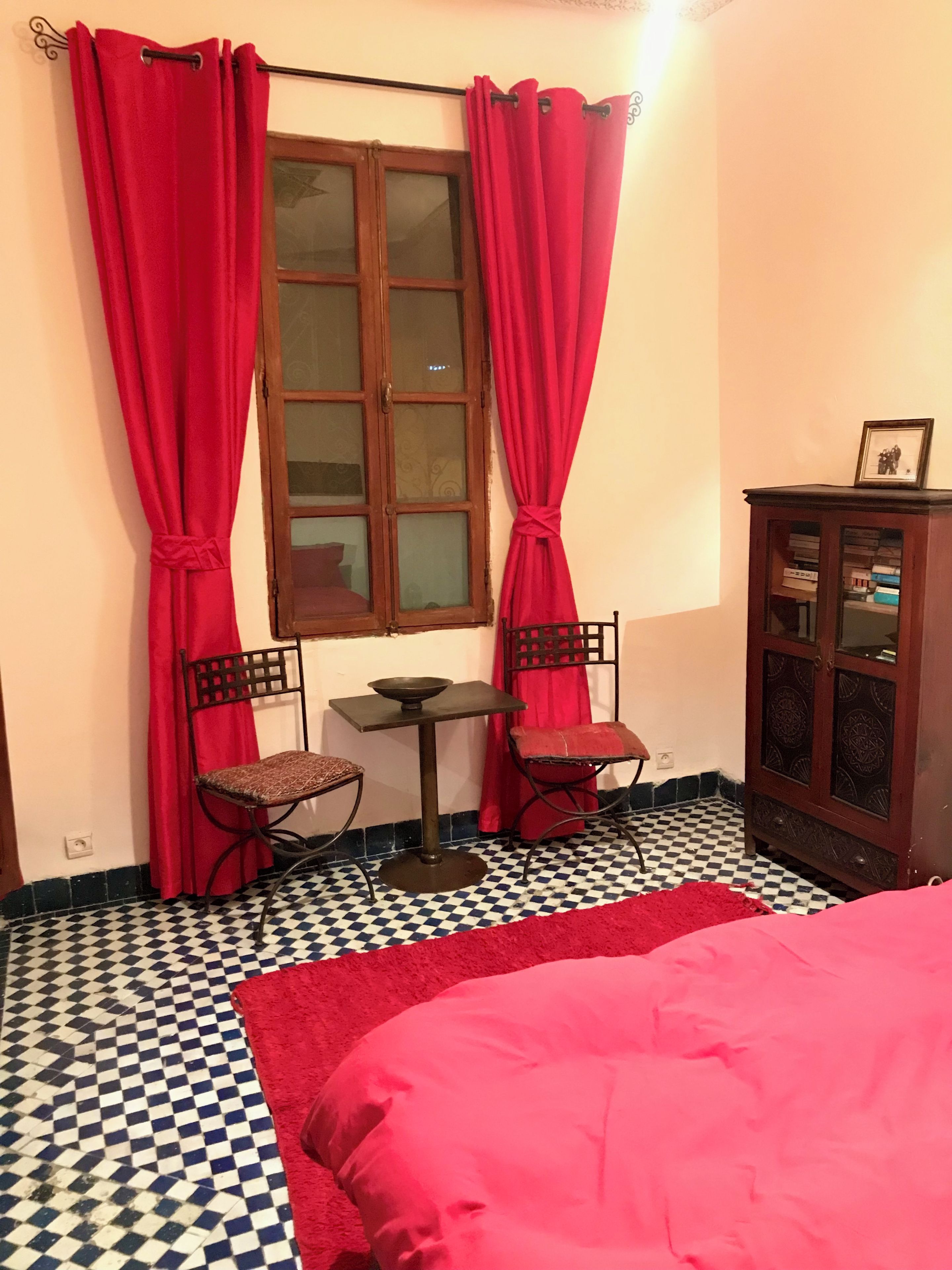 Une pièce avec un sol à carreaux, un lit recouvert d’une couverture rouge, deux chaises, une petite table, un meuble contenant des livres et une fenêtre avec des rideaux rouges.