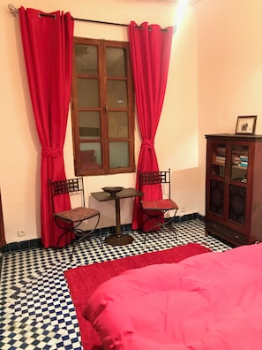 Une pièce avec un sol à carreaux, un lit recouvert d’une couverture rouge, deux chaises, une petite table, un meuble contenant des livres et une fenêtre avec des rideaux rouges.