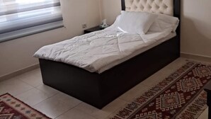 A bedroom with a bed, a nightstand, a window with blinds, and a patterned rug. - Spacious 3BR Amman Stay with Balcony & Free Parking (Amman)