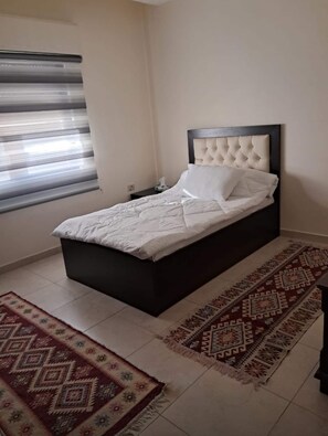 A bedroom with a bed, a nightstand, a window with blinds, and a patterned rug. - Spacious 3BR Amman Stay with Balcony & Free Parking (Amman)