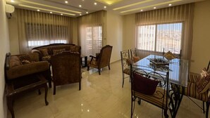 A modern living room with a sofa set, a dining table with chairs, and a large window with curtains. - Spacious 3BR Amman Stay with Balcony & Free Parking (Amman)