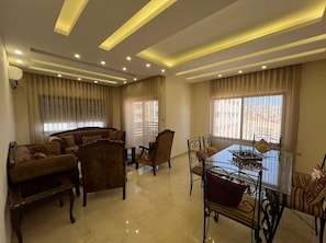 A modern living room with a sofa set, a dining table with chairs, and a large window with curtains. - Spacious 3BR Amman Stay with Balcony & Free Parking (Amman)
