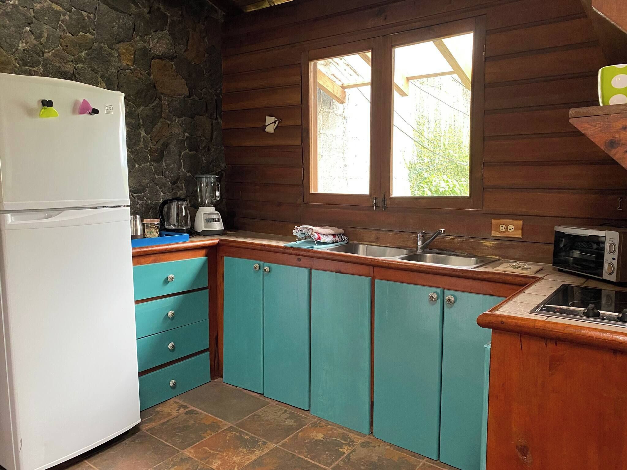 A compact kitchen with a stone wall, a white refrigerator, teal cabinets, a sink, and a window with a view.