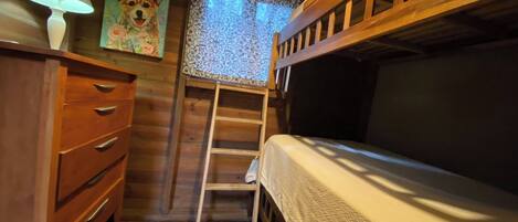 A wooden bunk bed room with a ladder, a desk with drawers, a lamp, and a painting of a dog.