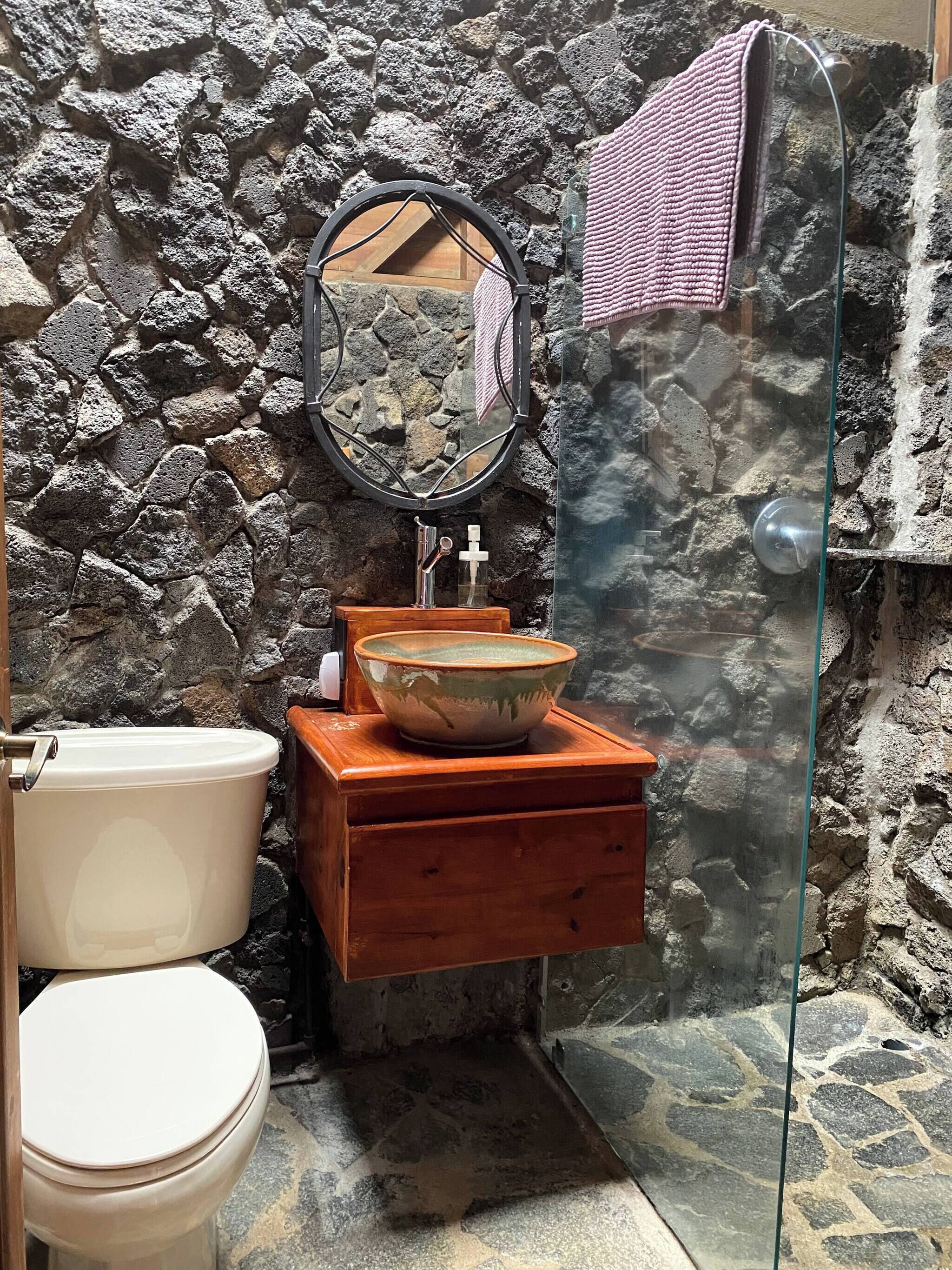 A bathroom with a stone wall, a wooden vanity, a glass shower enclosure, and a white toilet.