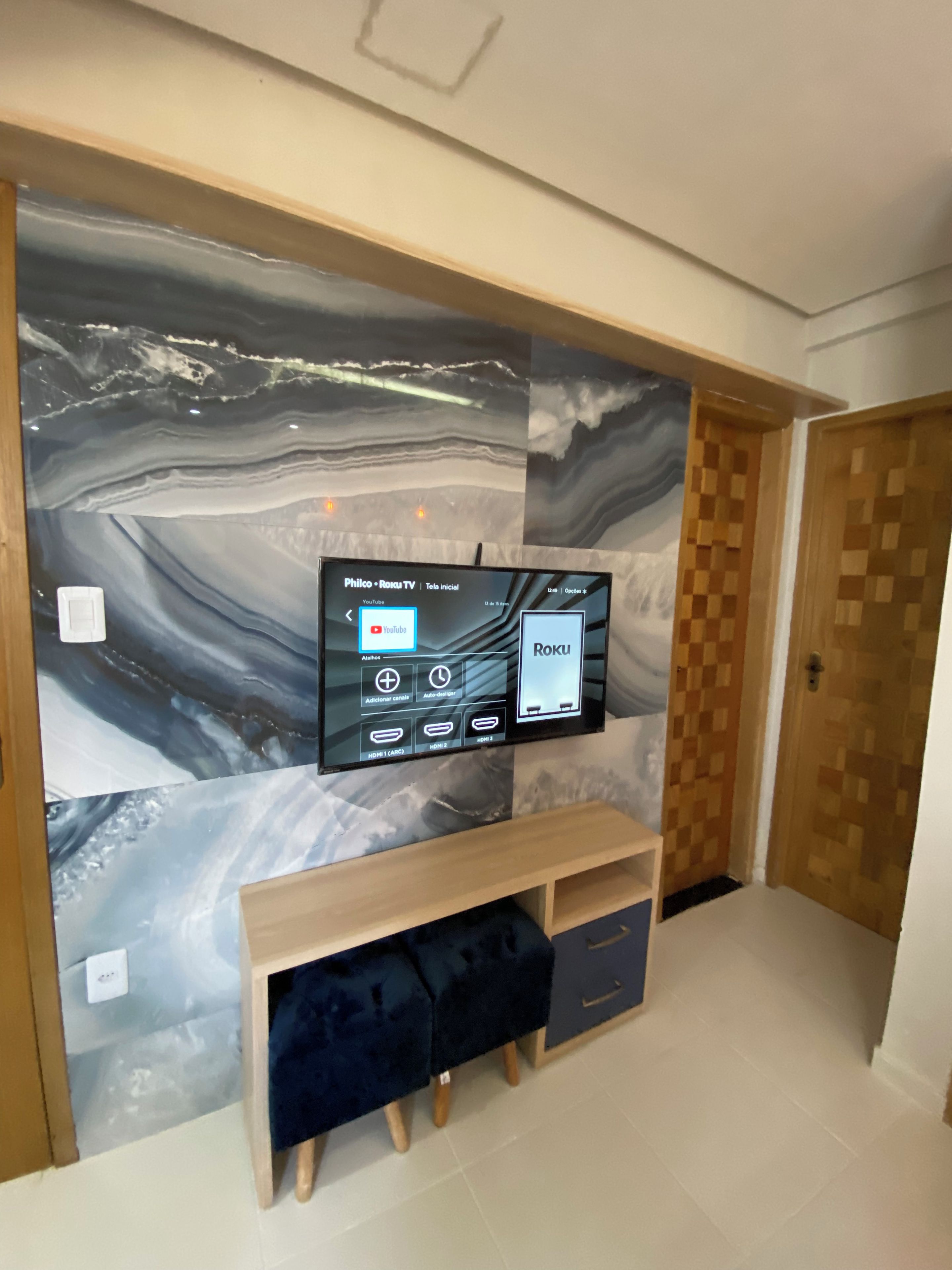 Un televisor de pantalla plana montado en una pared con diseño tipo mármol, un banco de madera debajo y una puerta a la derecha.