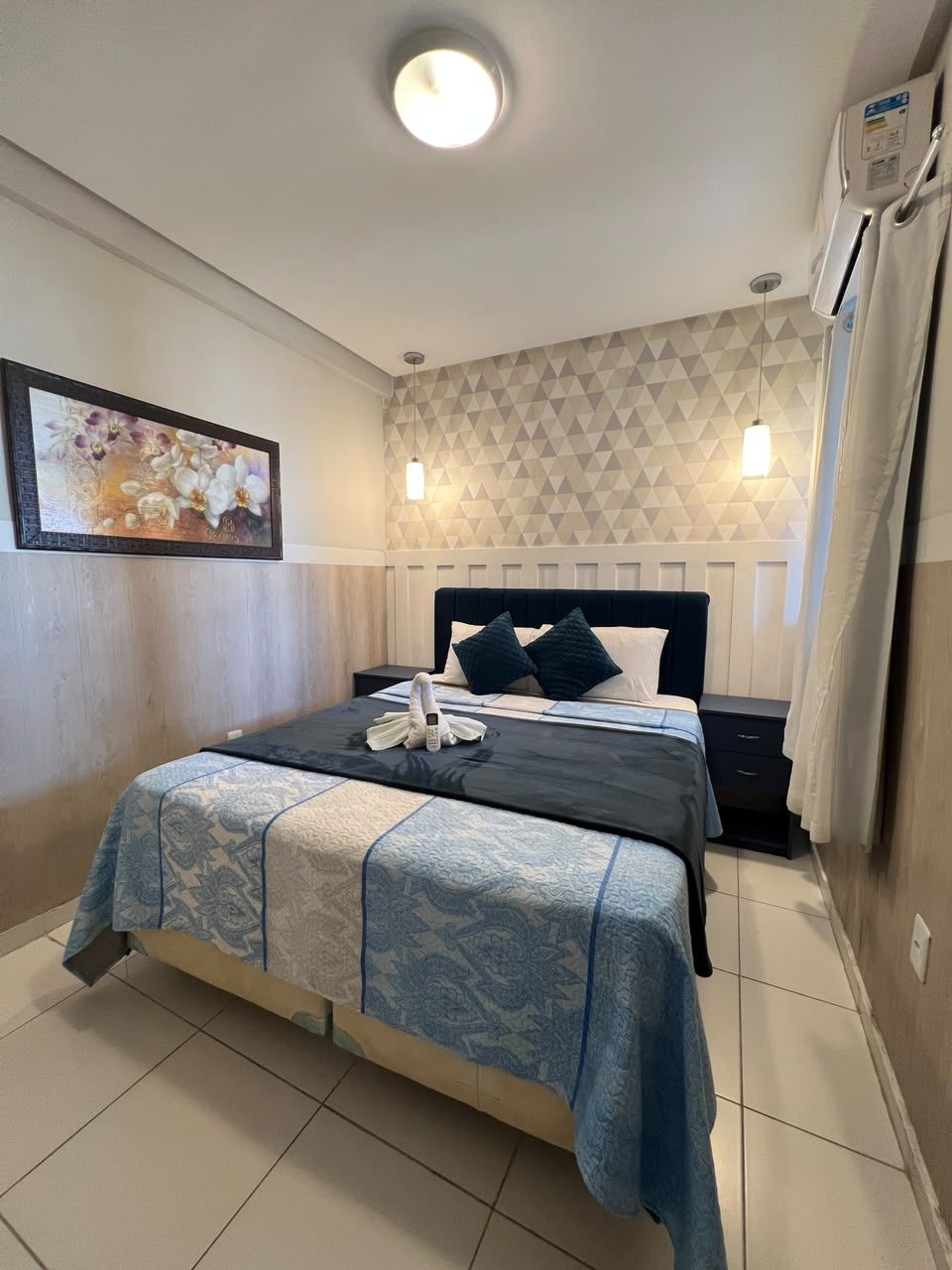 A hotel room with a bed, bedside table, and a wall-mounted air conditioning unit.