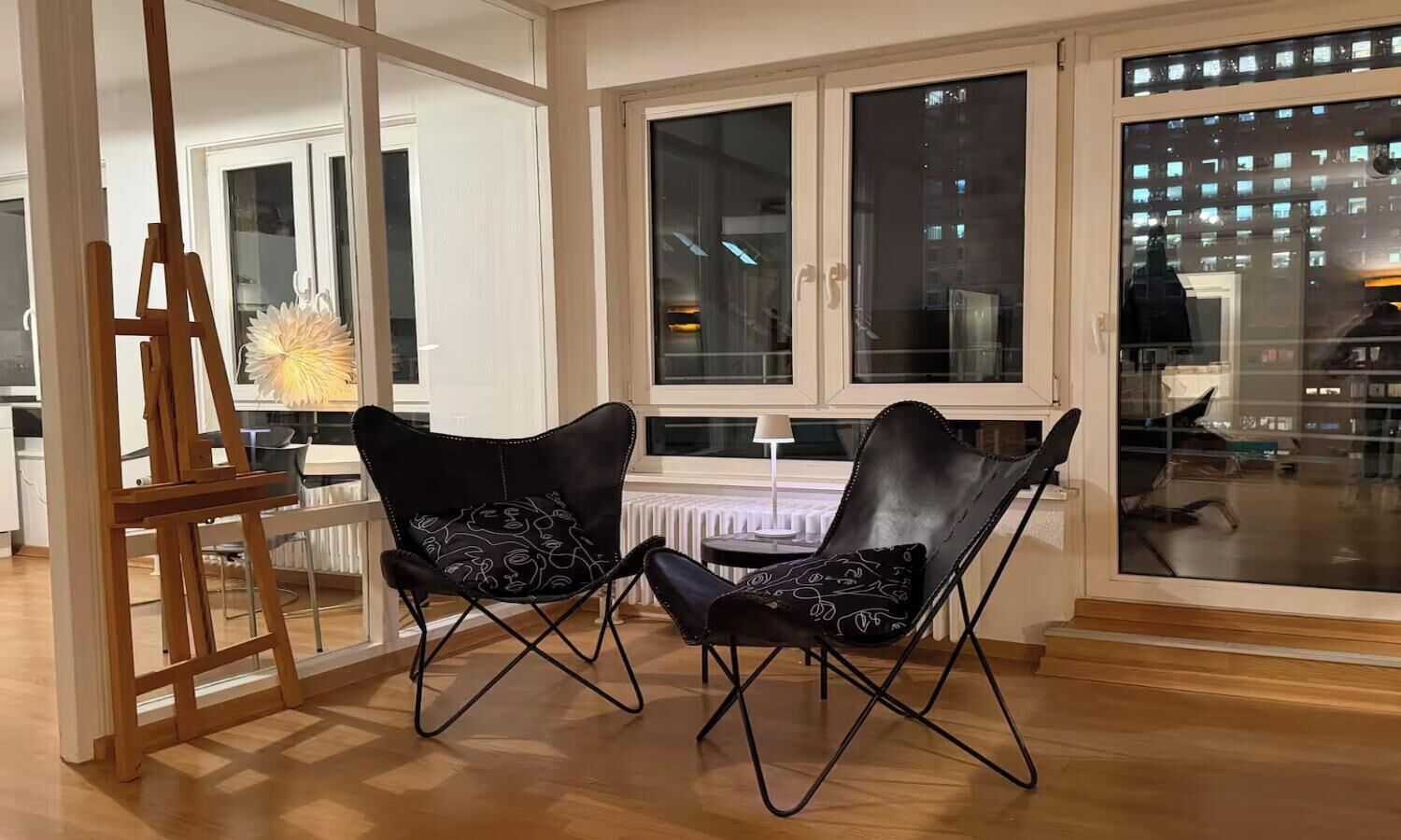 A room with a wooden floor, two black chairs with patterned cushions, a white radiator, and a window with a view of buildings.