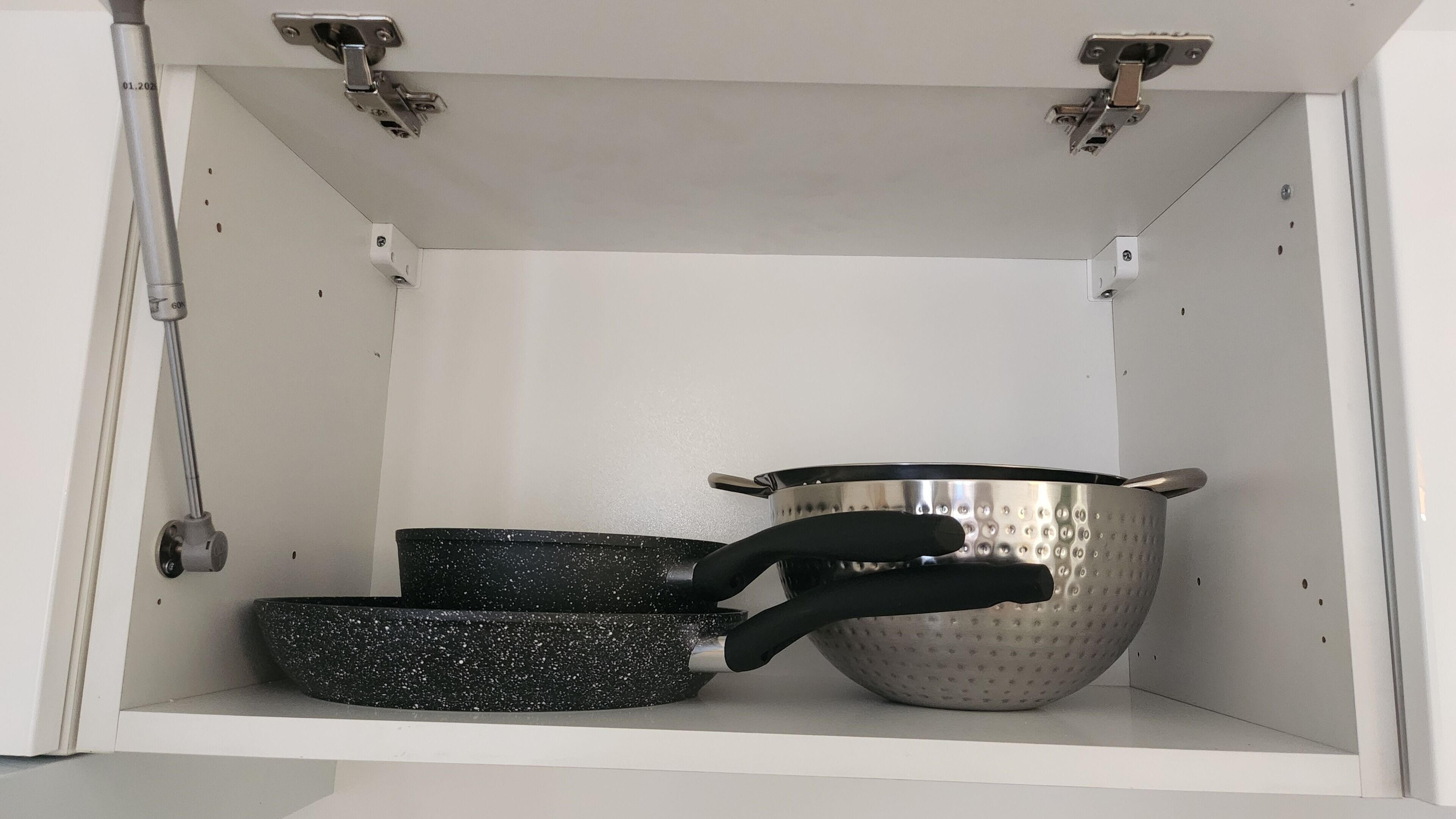 A kitchen cabinet with a metal pot, a frying pan, and a colander.