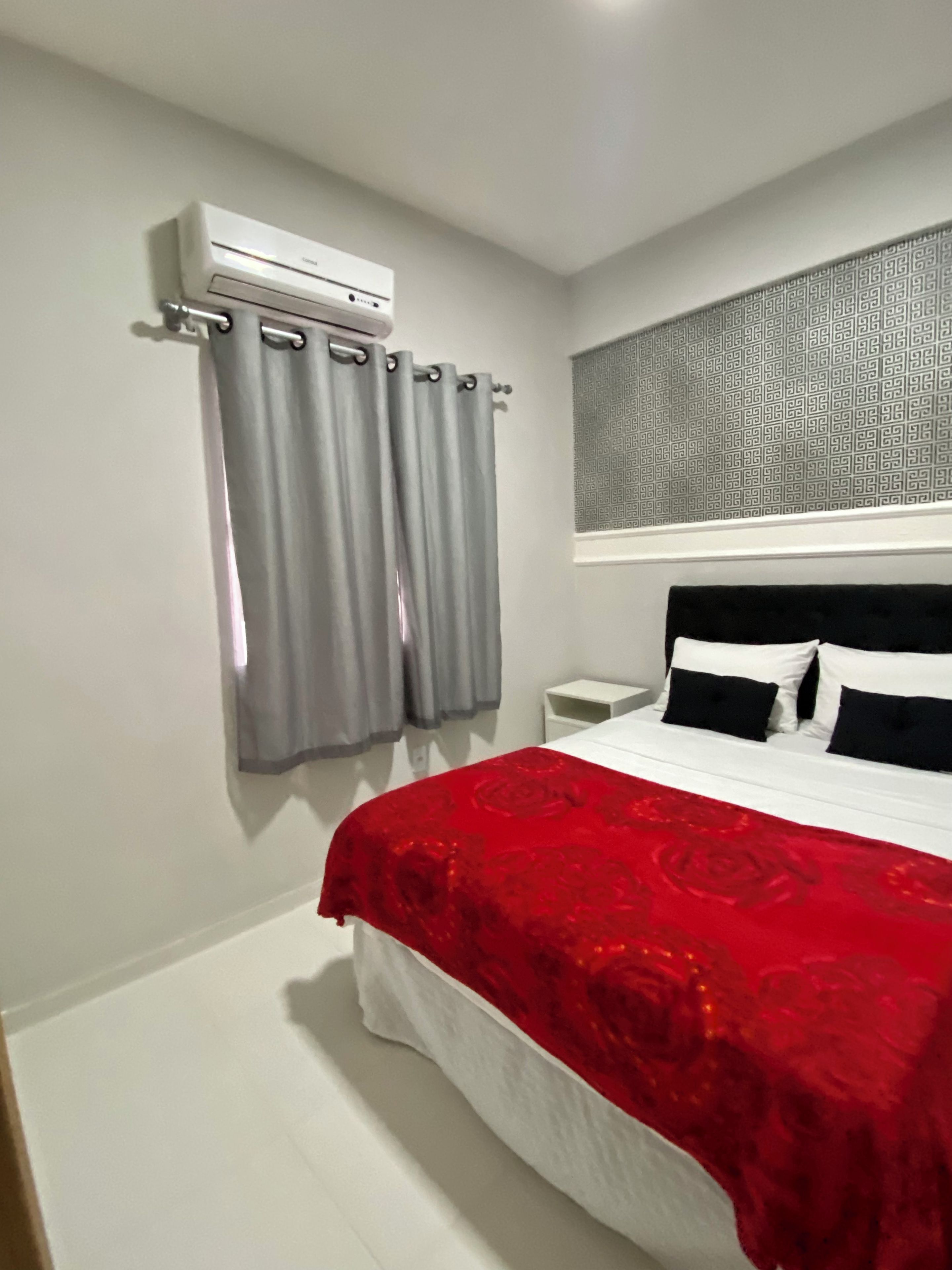 Une chambre à coucher comprenant un lit, une table de chevet, un appareil de climatisation et une fenêtre avec des rideaux à motifs.