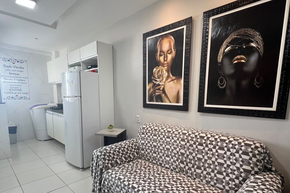 A modern living room with a patterned sofa, a kitchen area with white cabinets, and two framed artworks on the wall.