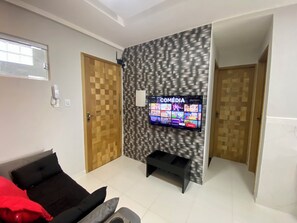Una sala de estar con un televisor de pantalla plana montado en una pared texturizada, una puerta de madera y un sofá negro con cojines rojos.