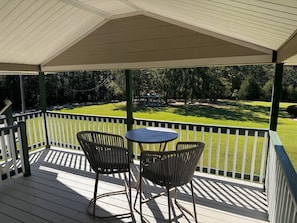 A covered outdoor area with a wooden deck, two chairs, a small round table, and a view of a manicured lawn and trees.