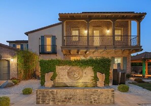 A two-story house with a stone facade, a balcony with a metal railing, and a stone fountain with a circular design in the center. - Spacious house with amazing view (West Covina)