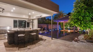 Een buitenkeuken met een bar en zitplaatsen, een zwembad met verlichting en een overdekt terras met een pergola.