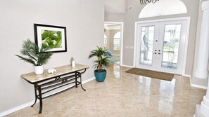 A hallway with a marble floor, a white bench, two potted plants, a framed picture, and a double door with glass panels. - Almare – Gulf Access, Pool, Spa & Boat Dock (Cape Coral)