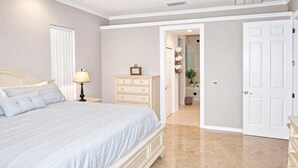 A bedroom with a bed, a nightstand, a dresser, and a door leading to a bathroom. - Almare – Gulf Access, Pool, Spa & Boat Dock (Cape Coral)