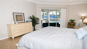 Ein Schlafzimmer mit einem großen Bett, einem Schminktisch, einem Spiegel, einer Topfpflanze und Blick auf einen Pool durch eine Schiebetür.