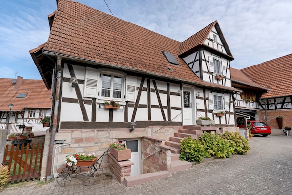 A traditional half-timbered house with a steeply pitched roof, a stone foundation, and a small front garden with a bicycle and flower pots.