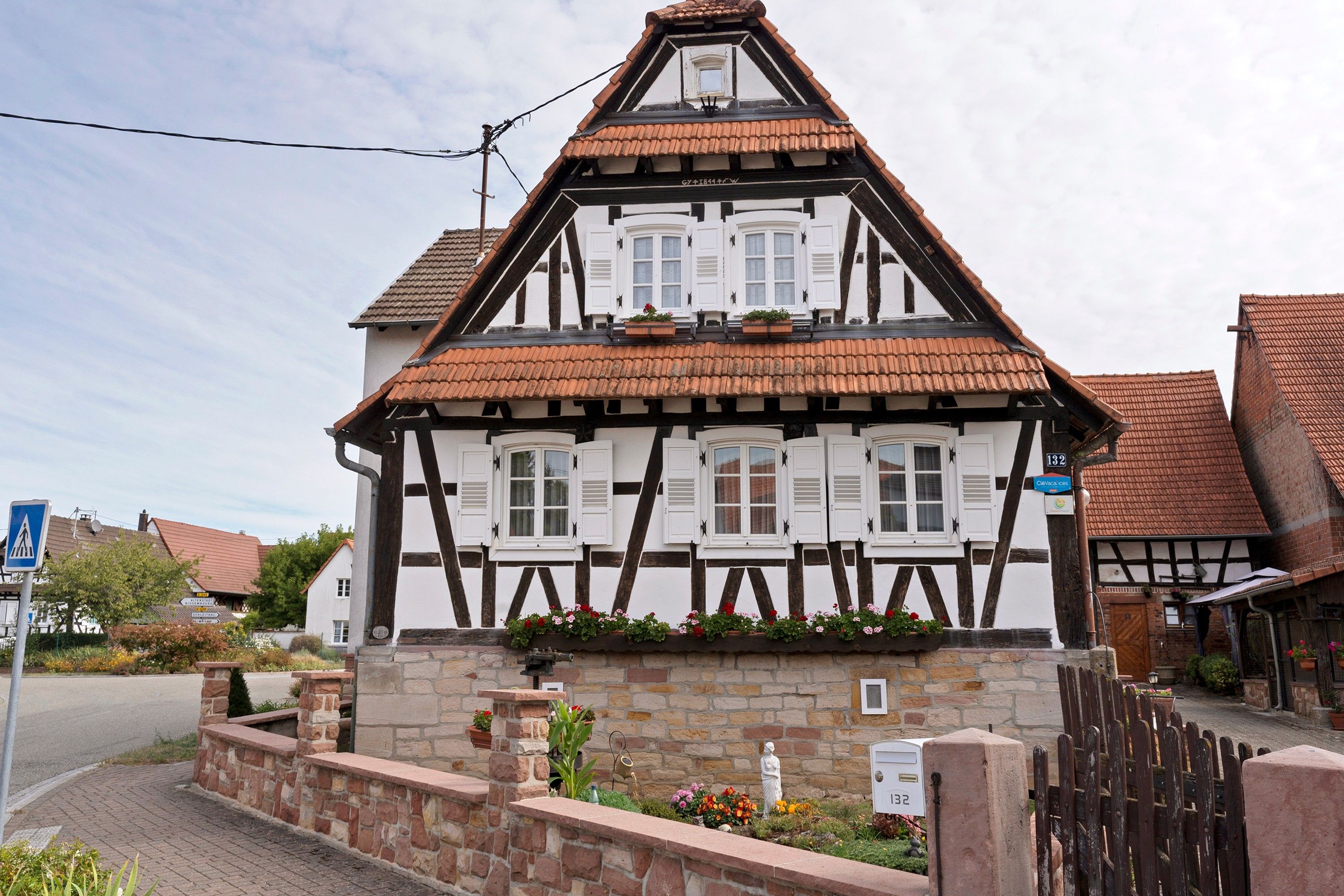 A traditional half-timbered house with a stone foundation, multiple windows with shutters, and a small garden area with flowers and a statue.