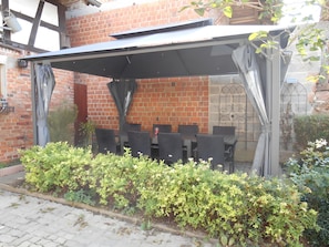 A covered outdoor seating area with a brick wall backdrop, surrounded by greenery.